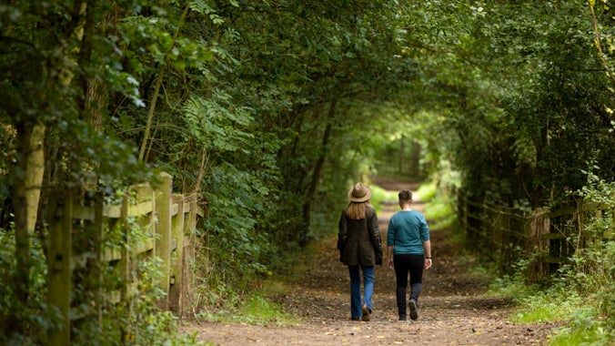 Image shows two women walking along a wide autumnal path with trees either side.